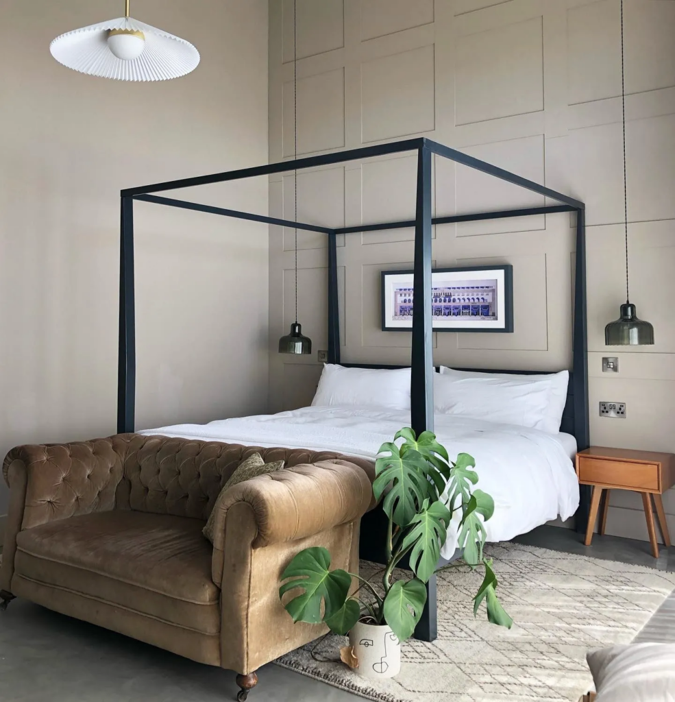 A modern bedroom with a black four-poster bed, white linens, a tufted brown loveseat, a monstera plant in a decorative pot, and pendant lights against a light brown panelled wall with Farrow & Bell.