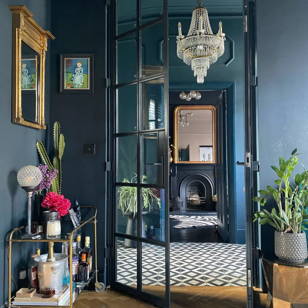 A stylish interior featuring Farrow & Ball Hague Blue and Black Blue paint, with a glass-paneled door leading to a richly decorated room with a black fireplace, a gold-framed mirror, and geometric floor tiles; the space is accented with a crystal chandelier, a gold bar cart with drinks and decor, and lush green plants.