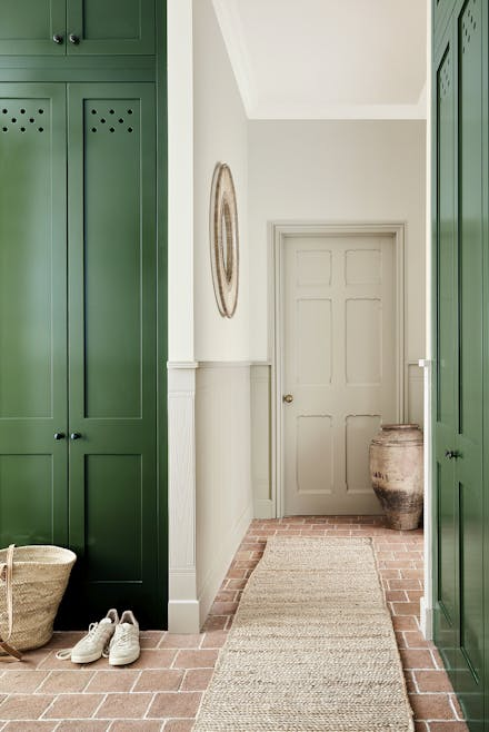 A bright and airy hallway with cabinetry painted in Little Greene Bushwhack Green, featuring natural textures such as a woven rug, terracotta floor tiles, and a straw basket, complemented by neutral walls, a beige door, and an earthy ceramic vase for a warm, inviting aesthetic.