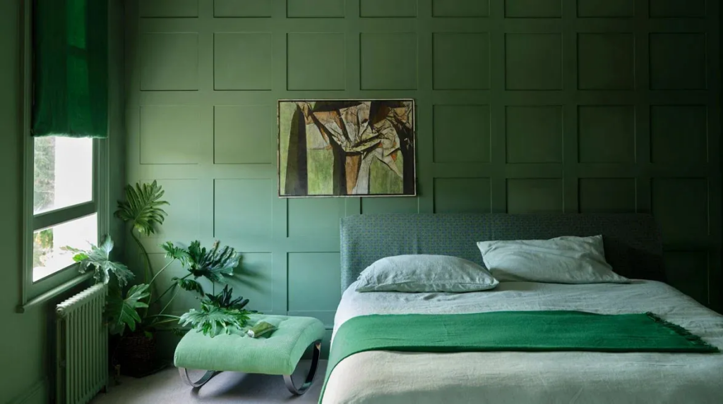 A tranquil bedroom featuring Farrow & Ball green panelled walls, a grey upholstered bed with white and green bedding, a matching green ottoman, lush indoor plants, and a large abstract painting, creating a harmonious, nature-inspired space.