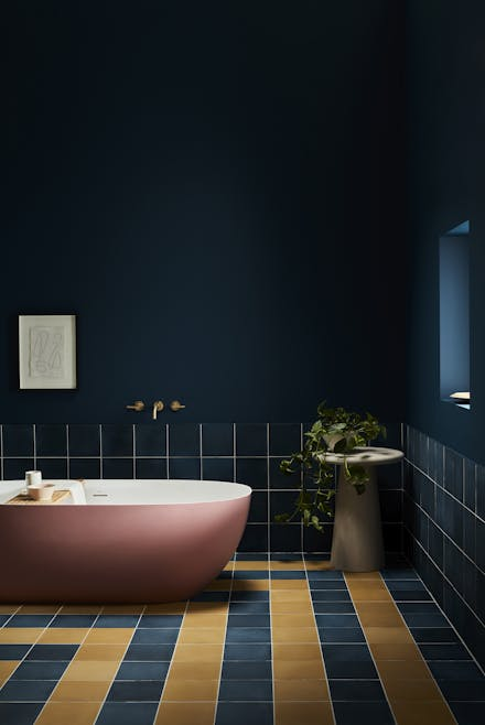 A stylish bathroom featuring walls painted in Little Greene Royal Navy, contrasted with a pink freestanding bathtub, navy and mustard floor tiles, and matching navy wall tiles; a sleek concrete pedestal sink with a trailing plant adds a touch of modern elegance.