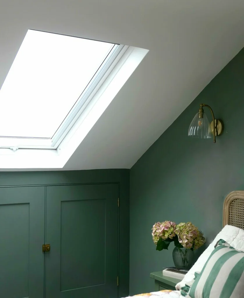 A cozy bedroom featuring walls and built-in storage painted in Farrow & Ball Calke Green, with a skylight allowing natural light to illuminate the space, a brass and glass wall sconce, a rattan headboard, and striped green and white bedding complemented by a vase of hydrangeas on the bedside table.