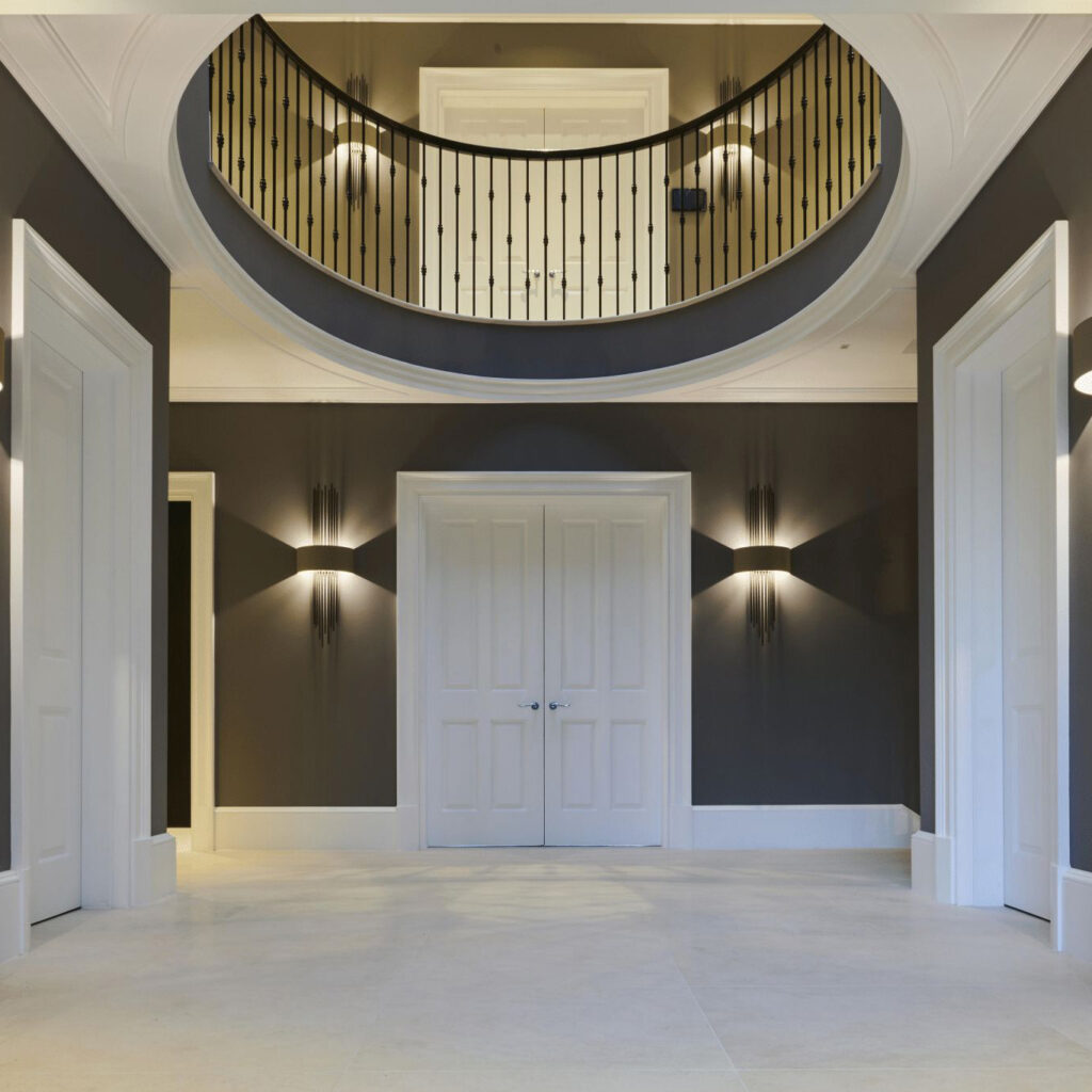 Monumental double-height entrance hall with curved balcony, panelled doors, and ambient wall lighting in a luxury London property.