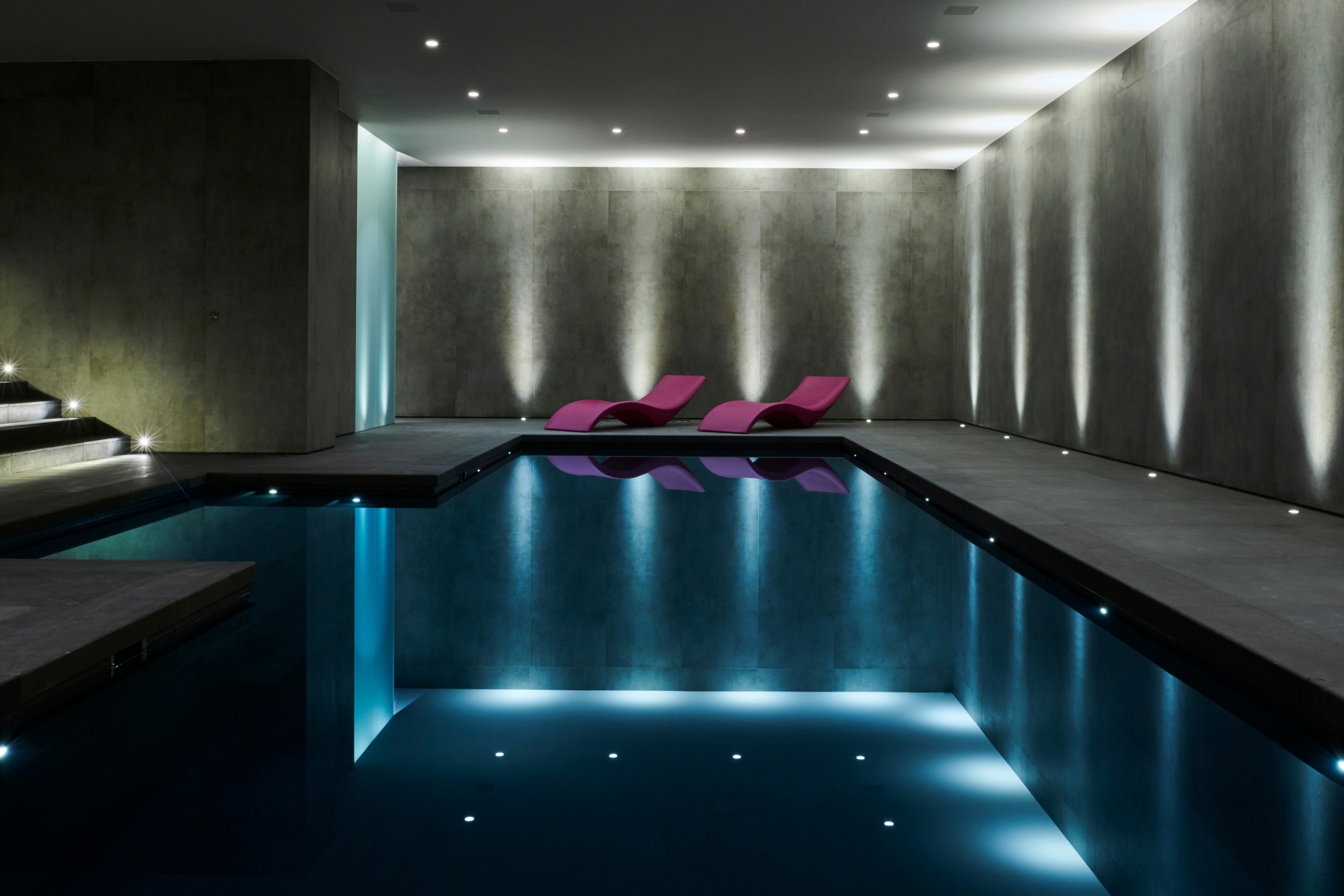 Minimalist indoor pool with moody lighting, concrete walls, and magenta lounge chairs.
